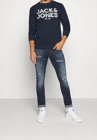 Man som bär en marinblå "JACK & JONES" sweatshirt, slitna slim-fit blå jeans med uppvikt nederkant, vita Adidas sneakers, står med en hand i fickan.
