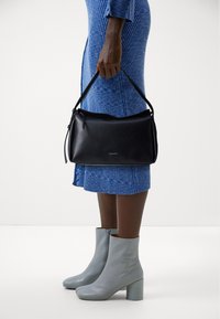Borsa a tracolla in pelle nera con chiusura a zip e forma arrotondata, tenuta da una persona che indossa un vestito blu a coste e stivali alla caviglia grigio chiaro.