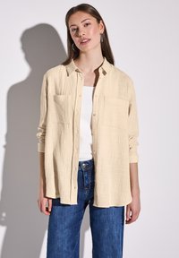 Béžová košile na knoflíky z texturované látky, s dvěma kapsami na hrudi, dlouhými rukávy s ohrnutými manžetami a volným střihem, v kombinaci s modrými džíny.