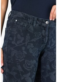 Tmavě modré džíny s květinovým vzorem, které mají klasický pětikapsový design, zapínání na knoflík a texturovanou látku.