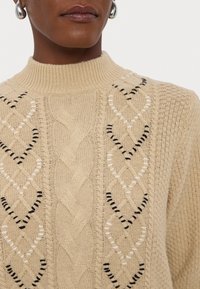 Bež narmatud särk kõrge kaeluse ja nöörmustrite ning must-valgete aktsentõmblustega. Struktuurne ja mugav välimus.