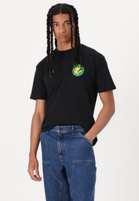 Black cotton t-shirt with short sleeves, featuring a green and yellow graphic logo on the left chest. Paired with blue denim jeans.