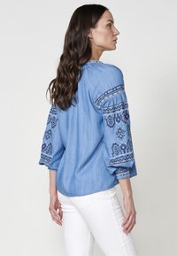 Ljus denimblus med broderade blå mönster på ärmarna, rundad nederkant, avslappnad passform och knäppta manschetter. Kombinerad med vita jeans.