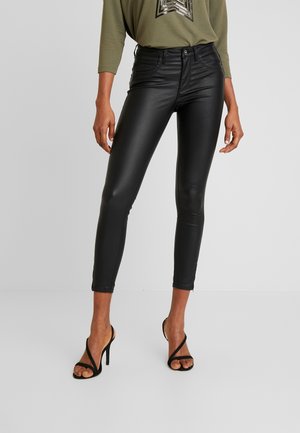 Personne portant un pantalon en cuir noir ajusté, un haut à manches longues vert olive et des sandales noires à talons hauts, debout devant un fond uni.