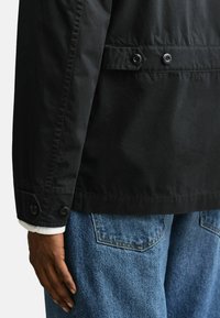 Nahaufnahme einer Hand, die neben einer blauen Jeans und einer schwarzen Jacke mit Knopfdetails an Manschette und Rücken ruht.