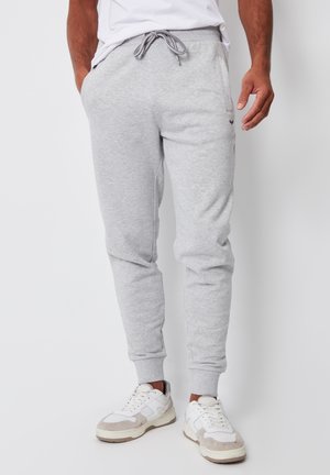 Mann trägt hellgraue Sweatpants mit Kordelzug und weiße Sneakers und steht vor weißem Hintergrund.