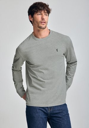 Gray long-sleeve t-shirt in soft fabric, featuring a small black logo on the chest. Worn with blue jeans. Casual fit and round neckline.