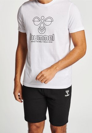 Mężczyzna noszący białą koszulkę hummel z logo i hasłem, w połączeniu z czarnymi szortami hummel z logo na lewym udzie.