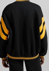 Svart sweatshirt med gula randiga detaljer på ärmar och krage. Har ribbade mudd och fåll; mjukt, texturerat tyg.