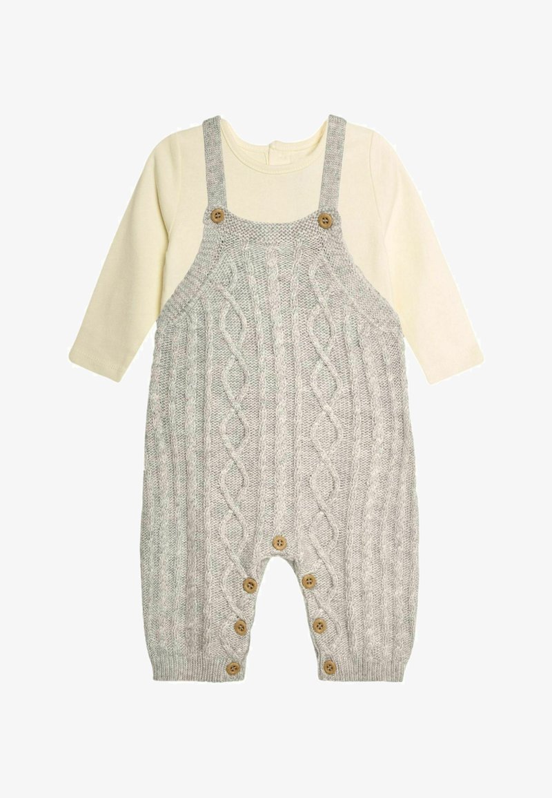 Szydełkowy szary kombinezon z wzorem w warkocze, ozdobiony drewnianymi guzikami i regulowanymi ramiączkami. Połączony z kremową bluzką z długim rękawem pod spodem.
