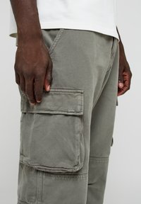 Pantalones cargo en verde oliva, con una tela texturizada. Incluyen bolsillos laterales y una etiqueta de marca discreta en el bolsillo inferior.
