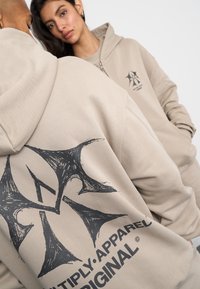 Felpa con cappuccio beige con un grande design grafico nero sul retro e testo del logo. Tessuto morbido con una vestibilità comoda e zip frontale.