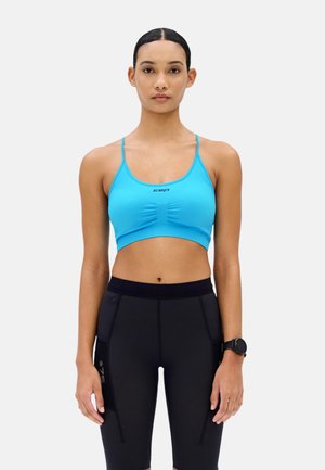 Woman wearing a blue sports bra and black high-waist athletic shorts with a smartwatch on her left wrist, standing against a plain background.