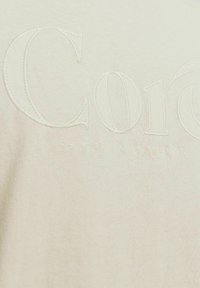 Kreemja värvi kangas, millel on broderitud tekst "Cor" ja koordinaadid "55.9427° N 9.1238° E" allpool kerge niidiga.