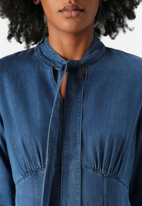 Denim blouse in dark blue with a keyhole neckline, tied collar, and gathered detailing at the waist. Smooth texture with subtle stitching.