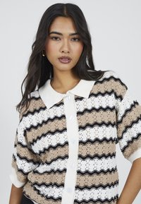 Sweter z dzianiny z falistym, paskowanym wzorem w odcieniach beżu, bieli i czerni. Z kołnierzykiem, dużymi guzikami i teksturowanym materiałem.