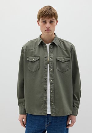 Camicia di cotone verde con maniche lunghe, chiusura con bottoni, due tasche sul petto e un taglio rilassato, indossata sopra una maglietta bianca e jeans blu.