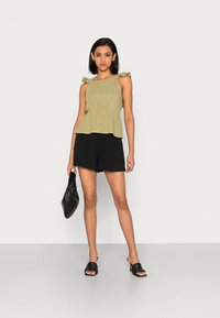 Top peplum verde oliva com alças de ombro com babados, combinado com calções pretos e uma bolsa preta de woven. O look é composto por materiais simples.