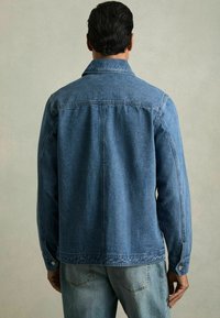 Jachetă din denim, albastru mediu, cu mansete cu buton, croială dreaptă, cusături vizibile, guler ascuțit, croiala lejeră, finisaj texturat subtil, vedere din spate.