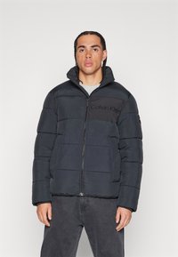 Juodas pūkų paltas su aukštu apykakle ir užtrauktuku. Tekstūruota medžiaga su kontrastinga juosta, ant kurios matomas "Calvin Klein" logotipas.