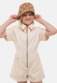 Enfant portant une combinaison beige à manches courtes avec fermeture éclair à l'avant et un chapeau de seau à pois bruns, tenant les côtés du chapeau, yeux plissés, souriant.