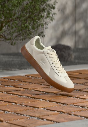 Biały niski sneaker z gumową podeszwą i wytłoczonym logo krokodyla unoszący się nad wzorzystą drewnianą podłogą na zewnątrz.