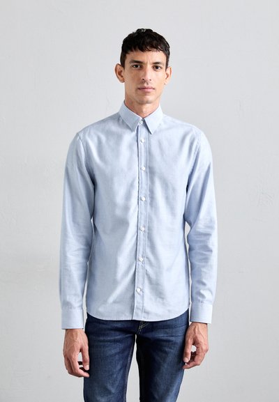 Camicia azzurro chiaro con colletto classico, maniche lunghe e bottoni bianchi, abbinata a jeans scuri, su uno sfondo neutro.