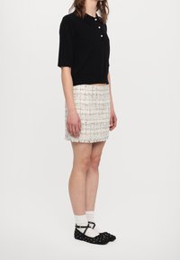 Camiseta polo negra de manga corta con tres botones, combinada con una mini falda blanca texturizada. El conjunto incluye zapatos planos negros con lunares y calcetines blancos.