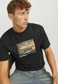 Sort bomulds t-shirt med et trykt billede af et værelse og ordet "Later." i hvid tekst. Korte ærmer, rounds halsdesign.