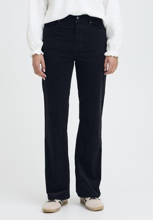 Person trägt schwarze Hose mit geradem Bein, weißen Langarm-Top mit Rüschenmanschetten und beige Sneaker mit schwarzen Streifen.