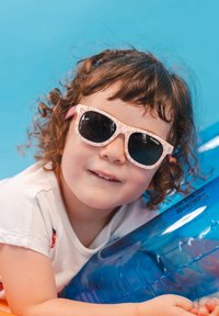 Occhiali da sole con montatura rosa a fantasia e lenti scure, indossati da un bambino. Sullo sfondo si vede un giocattolo gonfiabile blu per la piscina.