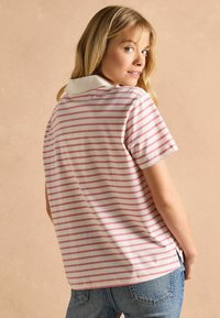 Īsām piedurknēm polo krekls baltā krāsā ar rozā svītrām, ar kontrastējošu baltu apkakli un brīvu griezumu. Tūlītējā izskatu papildina džinsu bikses.