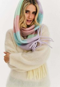 Blondynka nosząca pastelowy, wielokolorowy szalik na głowie i wokół szyi, ubrana w kremowy, puszysty sweter, z założonymi skrzyżowanymi ramionami.