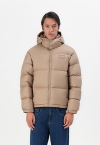 Beige pufferjakke med høj krave og hætte, med en frontlynlås, trykknapper og et logo på brystet. Båret over blå jeans.