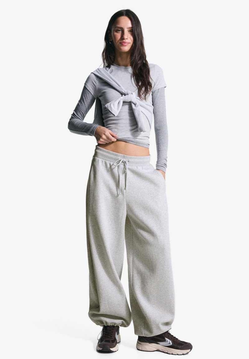 Kvinde i grå langærmet skjorte med bundet sweatshirt, løse grå sweatpants og sorte sneakers står med den ene hånd i lommen.