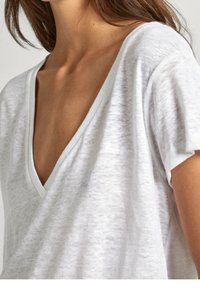 Žena v bílém, poloprůhledném tričku s krátkým rukávem a výstřihem do V, ukazujícím část krku a horní části hrudníku, s viditelnými hnědými vlasy.