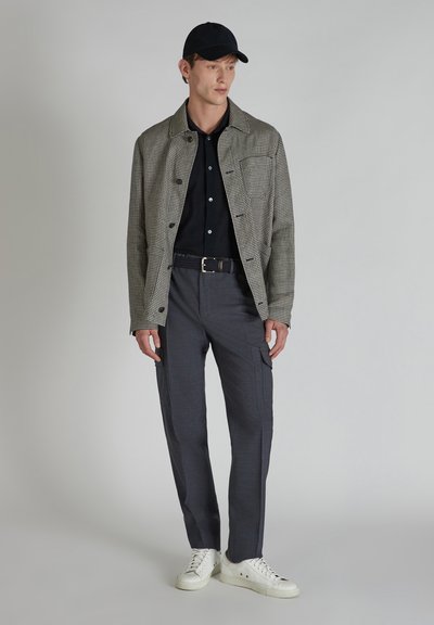 Parelgrijze jack over een zwart shirt, grijze cargobroek en witte sneakers. Het model draagt een zwarte pet en staat tegen een neutrale achtergrond.