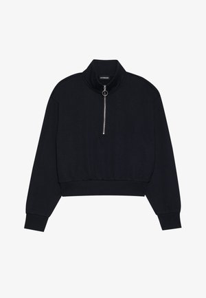 Melnā īsā puloverī, kas izgatavots no mīkstas auduma, ar augstu apkakli, priekšējo rāvējslēdzēju ar sudraba rokturi un ribotām piedurknēm un apakšmalu.