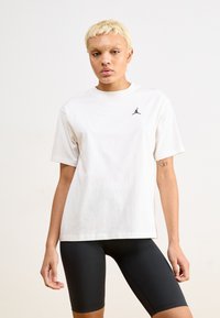 Hvit bomulls T-skjorte med kort erm, med en liten svart Jordan-logo på brystet. Kombinert med svarte sykkelshorts.