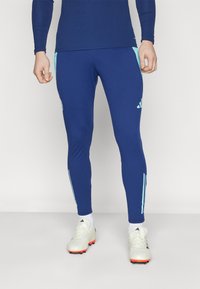 Pantaloni sportivi blu con accenti azzurri e design a tre strisce, realizzati in materiale elastico, stile attillato, abbinati a scarpini da calcio bianchi.