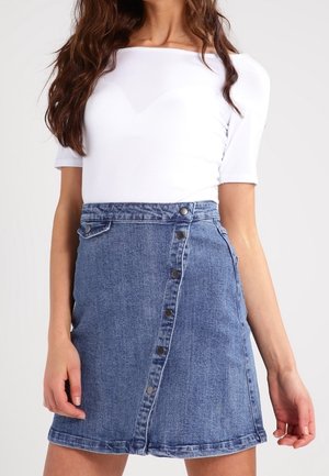 Kvinde iført en hvid kortærmet top og en blå denimnederdel med knapper foran, stående mod en ensfarvet lys baggrund.