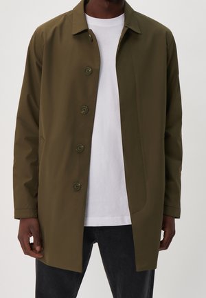 Mann, der einen olivgrünen knöpfbaren Mantel über einem weißen T-Shirt und schwarzen Hosen trägt, steht vor einem einfachen hellen Hintergrund.