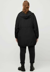 Svart lång hoodiejacka med elastisk fåll, i polyestermaterial, och sidofickor, kombinerad med svarta uppvikt byxor och kraftiga svarta kängor.