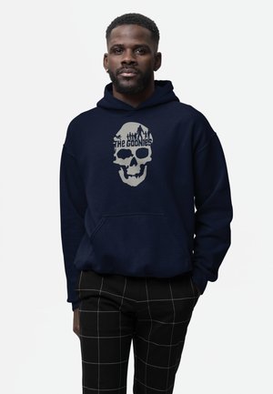 Man draagt een marineblauwe hoodie met "The Goonies" schedelgrafiek, gecombineerd met een zwarte broek met witte rasterlijnen, staand tegen een effen achtergrond.