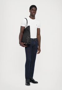 Bolso tote de cuero negro, camiseta blanca ajustada, jeans oscuros ajustados, zapatos negros. Modelo de pie posando con un brazo sosteniendo el bolso.