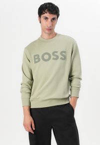 Sudadera verde clara con el logo "BOSS" en verde más oscuro. Presenta un cuello redondo, puños acanalados y un ajuste relajado. Usada con pantalones negros.