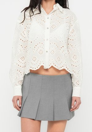 Kobieta ubrana w białą koronkową bluzkę na guziki z koronką typu eyelet i falowanym zakończeniem oraz szarą plisowaną mini spódnicę.
