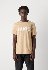 Šviesiai ruda medvilninė marškinėliai su ryškiu baltu užrašu "HUGO" ant priekinės dalies. Trumpomis rankovėmis ir apvaliu kaklu. Dėvimi su tamsiomis kelnėmis.
