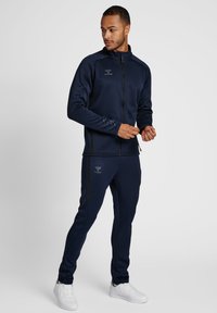 Ensemble de survêtement bleu marine en tissu doux, comprenant une veste zippée et un pantalon à taille élastique. Détail du logo sur les deux pièces. Baskets blanches.