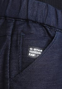 Ciemnoniebieska tkanina denimu o teksturowanej powierzchni, z małą kieszenią i metką z napisem "G-Star Originals RAW" w białym kolorze.